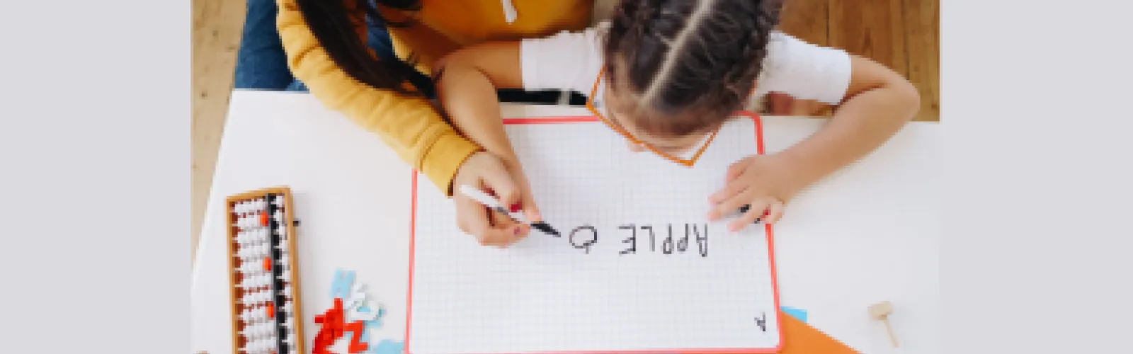 A girl writing the word apple on dry erase board on desk with adult hand guiding 
