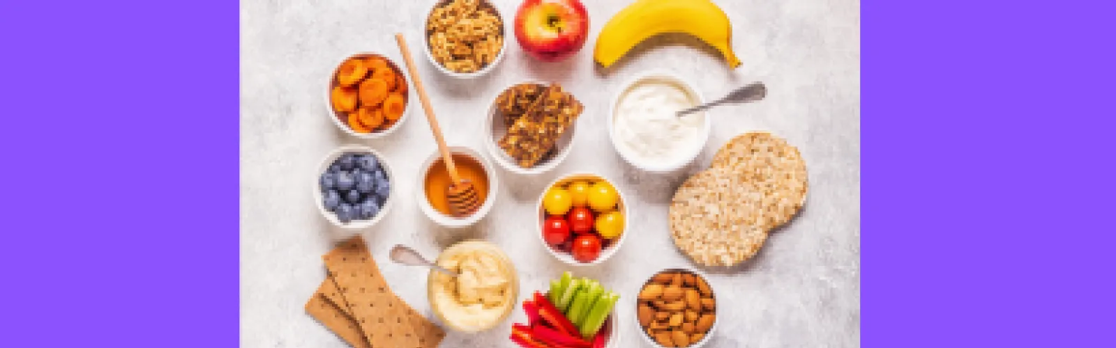 A variety of fruits, vegetables, and snacks artfully arranged on a purple background